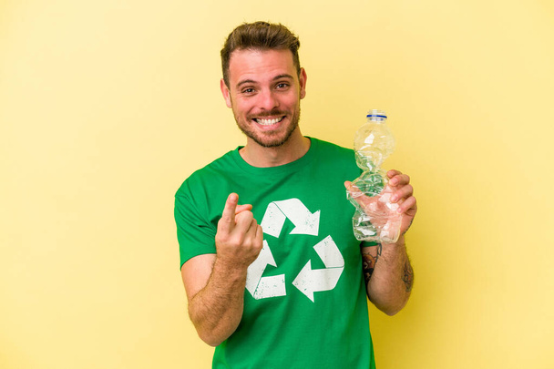 Young caucasian man holding a bottle of plastic to recycle isolated on yellow background pointing with finger at you as if inviting come closer. - Photo, Image