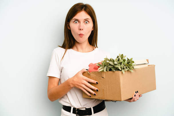 Giovane donna inglese fare una mossa, mentre raccogliendo una scatola piena di cose isolate su sfondo blu spalle spalle e occhi aperti confusi. - Foto, immagini