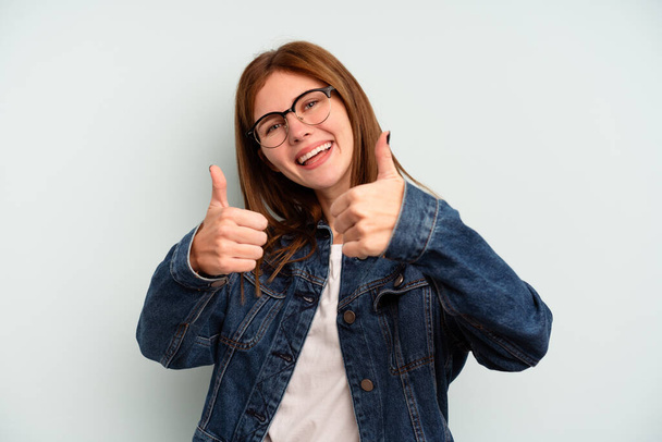 Jonge Engelse vrouw geïsoleerd op blauwe achtergrond het verhogen van beide duimen omhoog, glimlachen en zelfverzekerd. - Foto, afbeelding