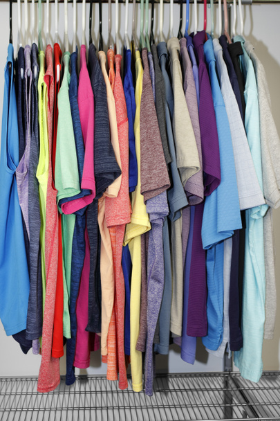 Hanging in a white wall closet are a variety of mens shirts including sleeveless, t shirts and polo shirts. Side view of mens sleeveless, t shirts and polo shirts hung on hangers in a closet closeup - Photo, Image