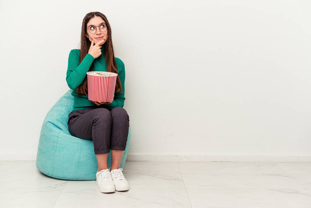 Junge kaukasische Frau isst Popcorn auf einem Puff isoliert auf weißem Hintergrund und schaut mit zweifelndem und skeptischem Gesichtsausdruck zur Seite. - Foto, Bild