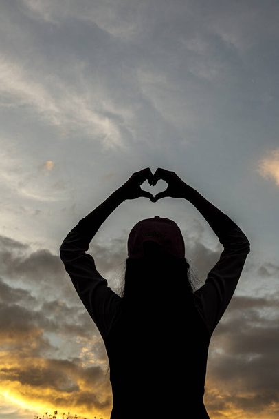 sylwetka młodej kobiety z podniesionymi rękami nad głową tworzy symbol w kształcie serca na pięknym tle nieba o zmierzchu. Koncepcja symbolu serca oznacza miłość, przyjaźń i dobroć. - Zdjęcie, obraz