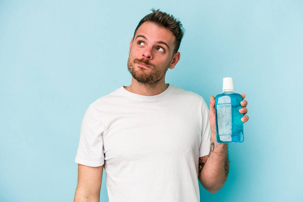 Young caucasian man holding mouthwash isolated on blue background dreaming of achieving goals and purposes - Photo, Image