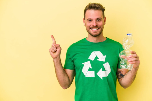 Mladý běloch drží láhev plastu na recyklaci izolované na žlutém pozadí, usmívá se a ukazuje stranou, ukazuje něco na prázdném místě. - Fotografie, Obrázek