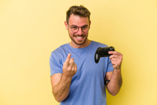 Jovem caucasiano segurando um controlador de jogo isolado no fundo amarelo apontando com o dedo para você como se convidando chegar mais perto. - Foto, Imagem