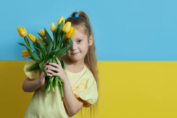 Cute child girl on the background of the Ukrainian flag holds a bouquet of yellow tulip flowers. yellow blue background - Фото, изображение