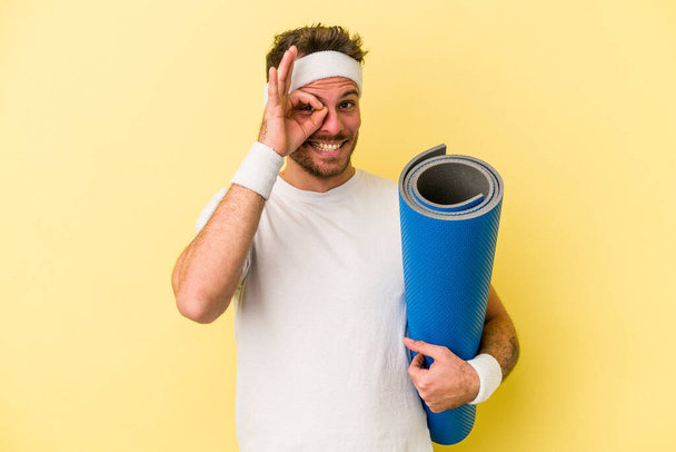Jeune homme caucasien sportif allant à des cours de yoga tout en tenant un tapis isolé sur fond jaune excité garder ok geste sur oeil. - Photo, image