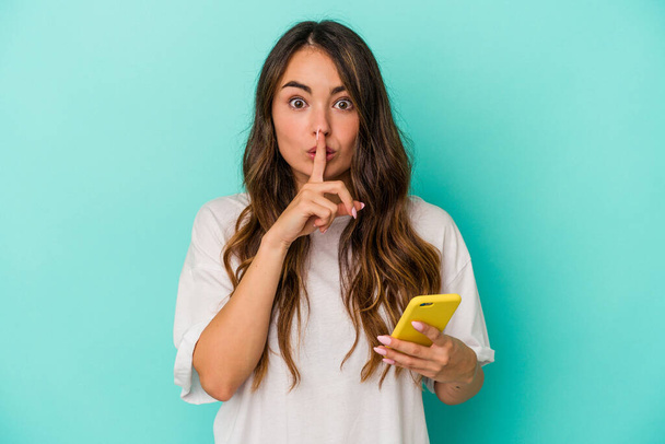 Young caucasian woman holding a mobile phone isolated on blue background keeping a secret or asking for silence. - Foto, immagini