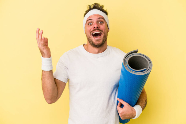 Молодий спортивний кавказький чоловік ходить на заняття з йоги, тримаючи килимок ізольований на жовтому тлі, отримуючи приємний сюрприз, збуджені та підняті руки
. - Фото, зображення