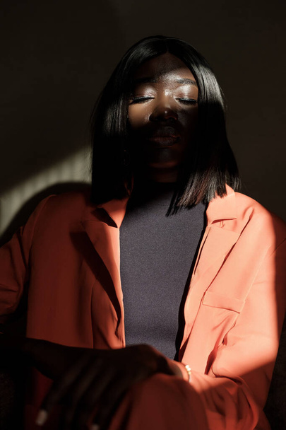 Czarna kobieta ma zamknięte oczy w ciemnym środowisku. Nosi pomarańczową kurtkę i jest w cieniu.. - Zdjęcie, obraz