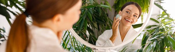Bornoz giyen mutlu genç bir kadın cildini silikon yüz fırçası kullanarak temizleme işleminden zevk alıyor, yeşil bitkilerle süslenmiş olarak banyoda duruyor. - Fotoğraf, Görsel