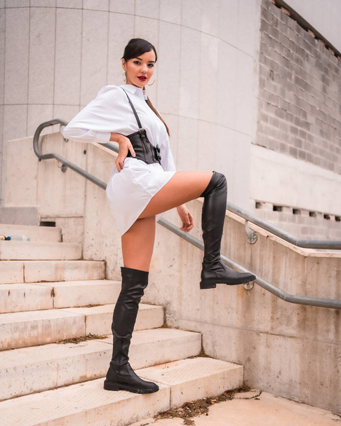 Young caucasian girl with white shirt and black leather boots. Enjoying the holidays in the city, going down the stairs with step signature - 写真・画像
