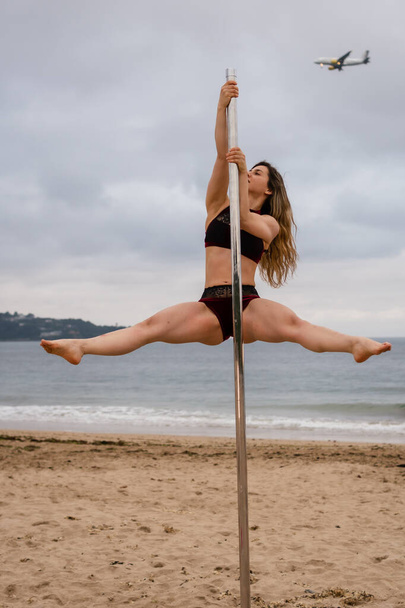Young caucasian girl performing pole dance at a beautiful beach. Acrobat performing pole dance in the sand of Hendaya beach, France. Dancing with an airplane in the background - Фото, зображення
