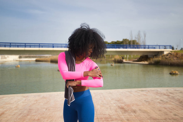 African-American woman with afro hair and sportswear, wearing fluorescent pink t-shirt and leggings, opening a running app on her cell phone. Fitness concept, sport, street, running. - Foto, imagen