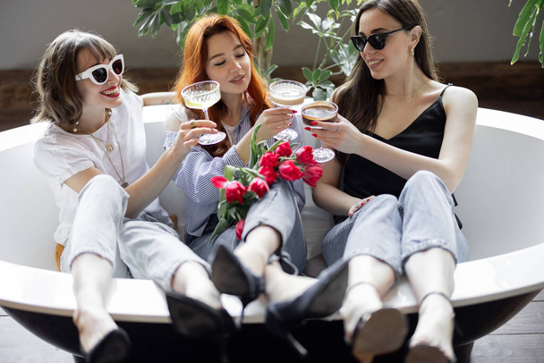 Drei junge erwachsene Freundinnen sitzen zusammen und trinken Alkohol in der Badewanne - Foto, Bild