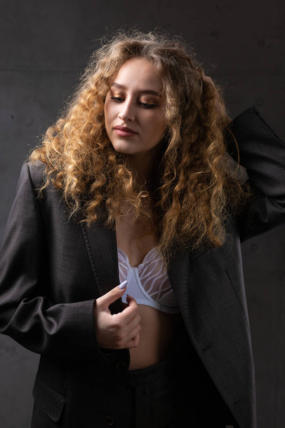 Ritratto di una giovane ragazza dai capelli rossi ricci sexy in una giacca con i capelli lunghi in studio. Foto drammatica in colori scuri. - Foto, immagini