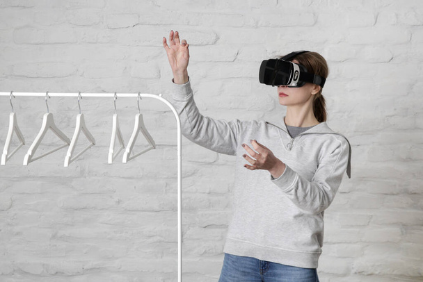 Portrait of woman with VR glasses choosing outfit from clothes hanger. Virtual reality experience and future technology concept.  - 写真・画像