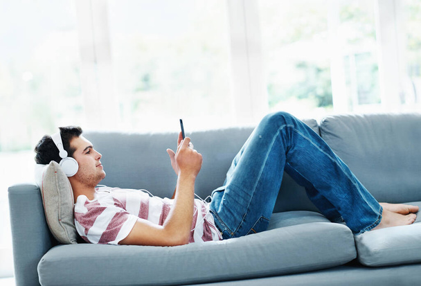 Spending time on his new tablet. An attractive man lying on the sofa at home and listening to music. - Fotoğraf, Görsel