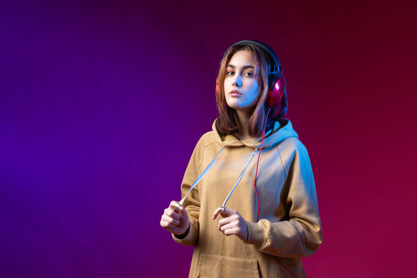 Young beautiful fashionable hipster girl dressed in a hoodie  listening to music in red headphones in a studio red dramatic background - Photo, image