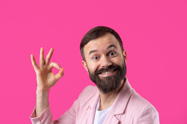 Πορτρέτο ενός χαμογελαστού νεαρού με ροζ μπουφάν. Άνθρωπος δείχνει ok χειρονομία, ενώ στέκεται στο στούντιο πάνω από απομονωμένο κόκκινο φόντο. - Φωτογραφία, εικόνα
