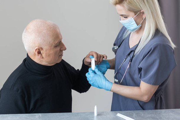 Close-up van medische staf handen introduceren van de nasale uitstrijkje aan de bufferbuis om een nasale cultuur monster te nemen van een senior man in zijn huis, op een huisbezoek. Snelle antigeentest op Covid-19 - Foto, afbeelding