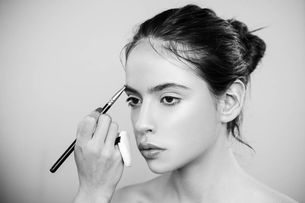 Beautiful young woman getting eyebrow make-up. The artist is applying eyeshadow on her eyebrow with brush. Young model girl getting her natural eyebrow makeup. - Foto, imagen