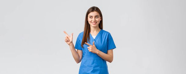 Travailleurs de la santé, prévenir le virus, concept d'assurance et de médecine. Jolie infirmière souriante ou médecin en blouse bleue, les doigts pointant vers la gauche et regardant baner avec sourire heureux, fond gris - Photo, image