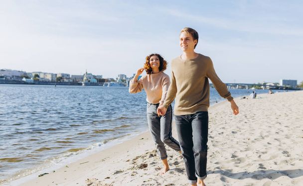 Δύο ερωτευμένοι νεαροί να τρέχουν στην παραλία κρατώντας τα χέρια. Χαμογελάνε ο ένας στον άλλο. Καλό ζευγάρι. Παραλία. Αισθήματα. ντυμένοι με πουλόβερ - Φωτογραφία, εικόνα