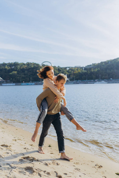 Kumsaldaki mutlu genç çift. Adam, aşık bir çiftin kumlu portresinin üzerinde duran kızı kollarında tutuyor. - Fotoğraf, Görsel
