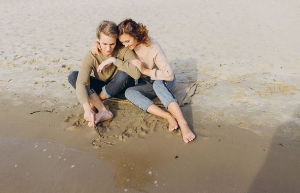 Kochająca się para bawi się - śmieją się, przytulają i cieszą ciepłym letnim wieczorem. Romantyczna para siedząca nad morzem. Męskie i żeńskie nogi na plaży. Facet rysuje na piasku. Serce - Zdjęcie, obraz