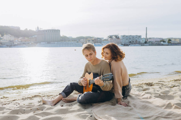 Ο άνθρωπος φλερτάρει παίζοντας κιθάρα ενώ ένα κορίτσι φαίνεται έκπληκτος με τη θάλασσα στο παρασκήνιο - Φωτογραφία, εικόνα