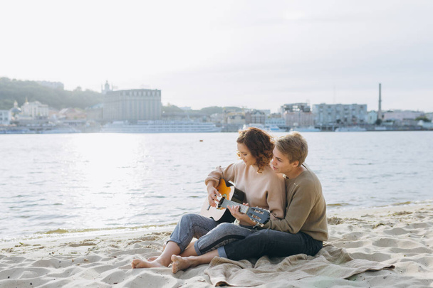 Genç adam kız arkadaşına gitar çalmayı öğretiyor, kendi kendine öğreniyor. Aşık bir çift kumsalda eğleniyor. - Fotoğraf, Görsel