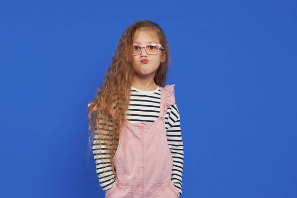 Portrait of a funny girl in glasses on a blue background. Child for an eye test. Stylish child holding glasses and looking at the camera. Vision measurement for schoolchildren. Back to school. - Fotó, kép