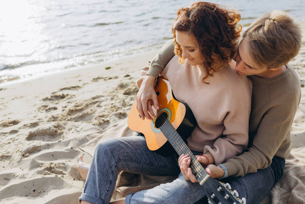 Młody facet uczy swoją dziewczynę gry na gitarze, samoukiem. Para zakochanych bawiących się na plaży - Zdjęcie, obraz