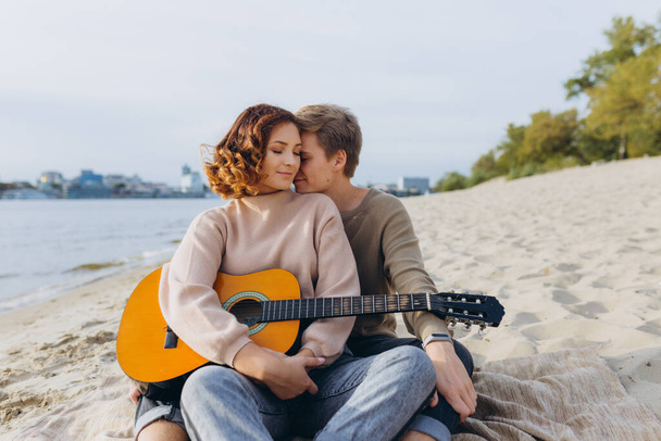 Genç adam kız arkadaşına gitar çalmayı öğretiyor, kendi kendine öğreniyor. Aşık çift kumsalda eğleniyor, sarılıyor ve öpüşüyor. - Fotoğraf, Görsel