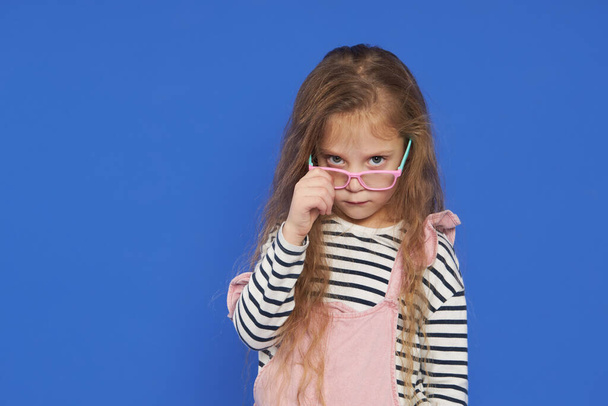 Portrait of a funny girl in glasses on a blue background. Child for an eye test. Stylish child holding glasses and looking at the camera. Vision measurement for schoolchildren. Back to school. - Foto, afbeelding