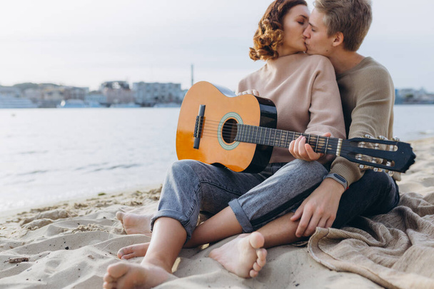 Молодой парень учит свою девушку играть на гитаре, самоучка. Пара влюбленных, развлекающихся на пляже, обнимающихся и целующихся - Фото, изображение
