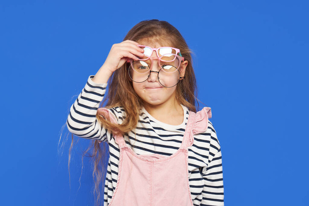 Portrait of a funny girl in glasses on a blue background. Child for an eye test. Stylish child holding glasses and looking at the camera. Vision measurement for schoolchildren. Back to school. - Foto, Imagen