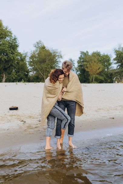 A loving couple has fun - they laugh, hug each other and enjoy a warm summer evening. Romantic couple standing by the sea. Wrapped in a warm blanket - Zdjęcie, obraz