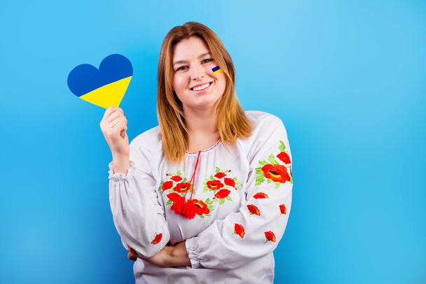 Uma jovem ucraniana com a bandeira da Ucrânia em seu rosto em uma camisa bordada segura um coração de uma bandeira amarelo-azul. O conceito de participação do povo ucraniano na guerra com a Rússia - Foto, Imagem