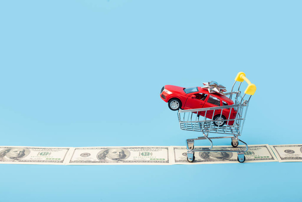 red toy car in shopping cart, on money road isolated on blue background - Фото, изображение