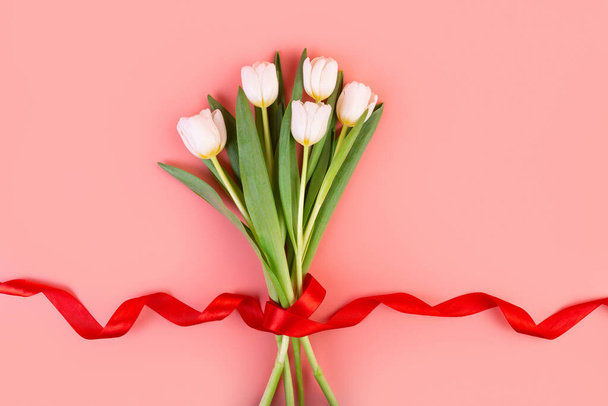 A virágok fehér tulipánok, lapos. Egy gyönyörű csokor piros szalaggal és egy pasztell rózsaszín háttéren elszigetelt borítékkal. tárgy dekoráció a nők napja, anyák napja, évforduló. - Fotó, kép
