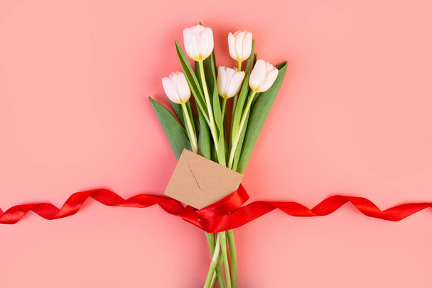 I fiori sono tulipani bianchi, piatti. Un bellissimo bouquet legato con un nastro rosso e una busta isolata su uno sfondo rosa pastello. oggetto per la decorazione per la festa della donna, festa della mamma, anniversario. - Foto, immagini