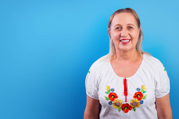 Eine Ukrainerin in einem bestickten Hemd lächelt glücklich isoliert auf blauem Hintergrund mit Kopierraum. Das Konzept der Beteiligung des ukrainischen Volkes am Krieg gegen Russland. Kein Kriegskonzept - Foto, Bild