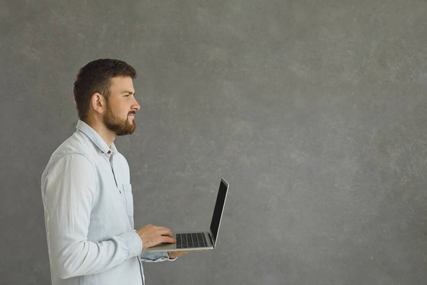 Portrait of a young and confident man standing on a gray background with a laptop in his hands. - Foto, Bild