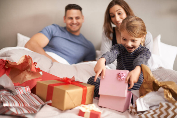 Oh wow, thanks mom and dad. Shot of a little girl opening presents in bed as her parents watch. - Photo, Image