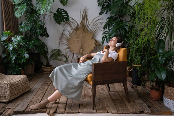 Calma fiorista donna in cuffie moderne ascoltare musica rilassarsi nel giardino di casa con diverse piante da casa - Foto, immagini