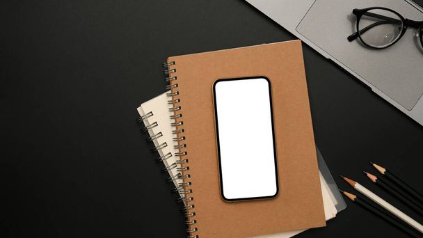 Overhead shot, Μοντέρνος χώρος εργασίας με λευκό smartphone οθόνη mockup πάνω από notebook, laptop και αξεσουάρ σε μαύρο φόντο. - Φωτογραφία, εικόνα