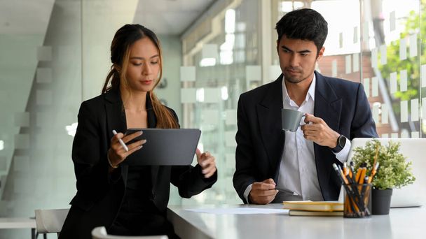 Smart Asian businessman concentrated working with female coworker, looking the data at tablet screen and drinking morning coffee. - Photo, Image