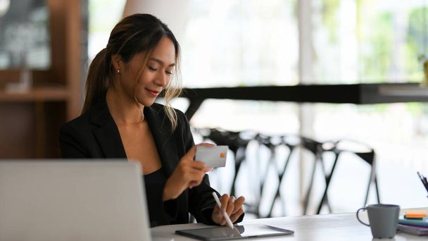 Χαρούμενη Ασιάτισσα επιχειρηματίας που κάνει online πληρωμές με πιστωτική κάρτα μέσω του υπολογιστή tablet στο γραφείο της. Online πληρωμή, μεταφορά χρημάτων. - Φωτογραφία, εικόνα